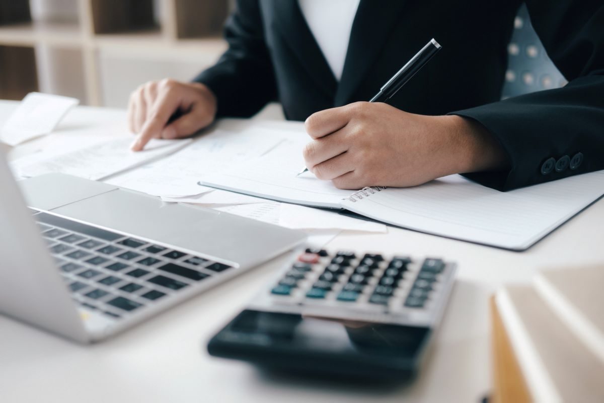 Person sat at a desk writing notes with a calculator and laptop on the table