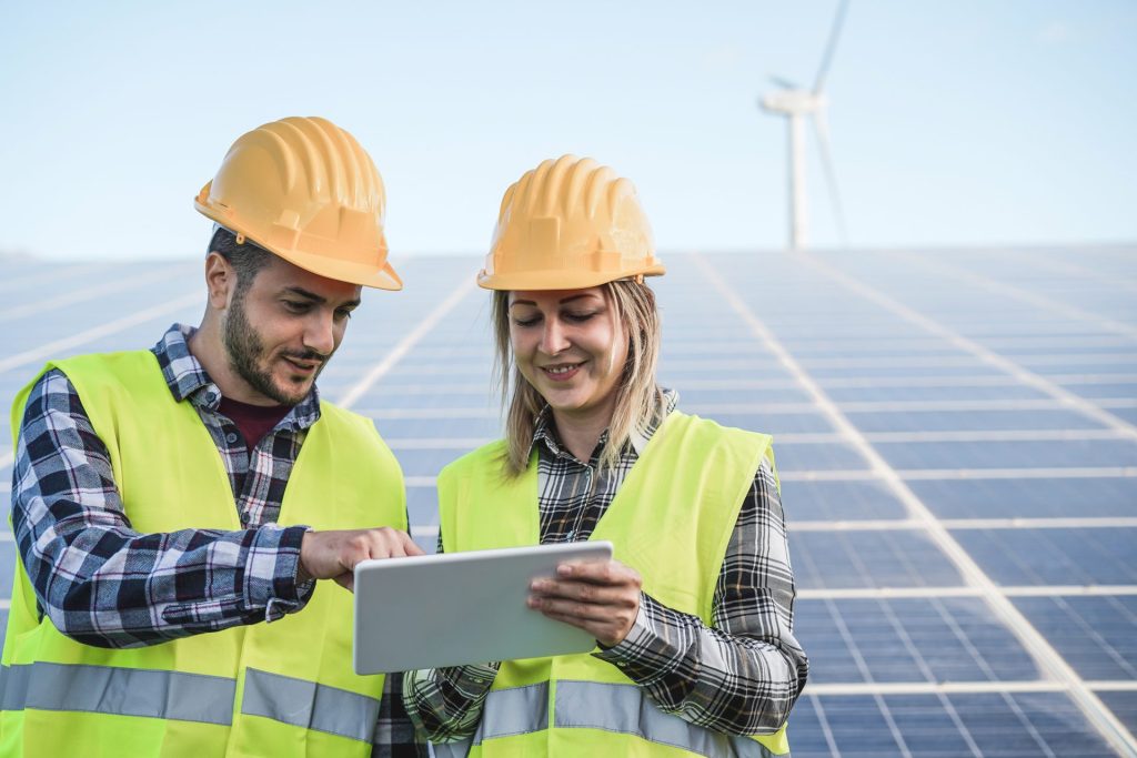 Two workers inspecting information on an iPad with solar panels and a wind turbine in the background