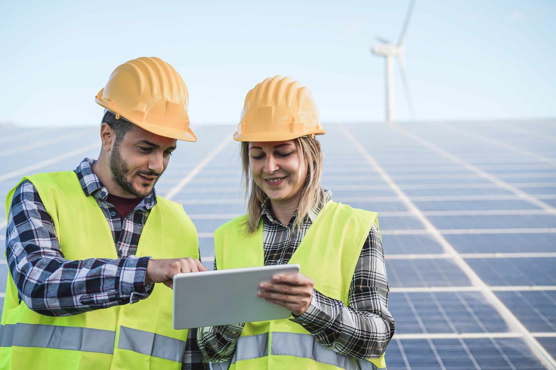 Two workers inspecting information on an iPad with solar panels and a wind turbine in the background