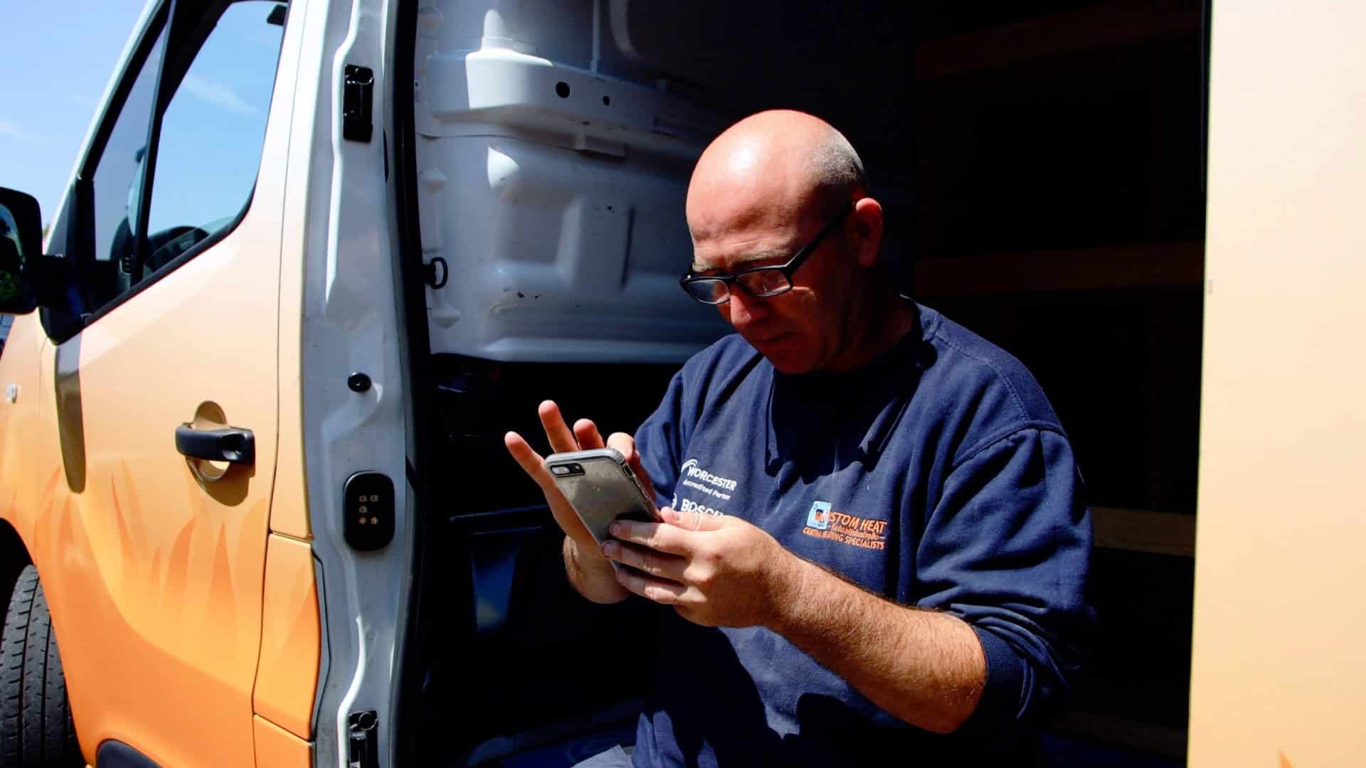 Custom Heat engineer sitting in van looking at mobile phone